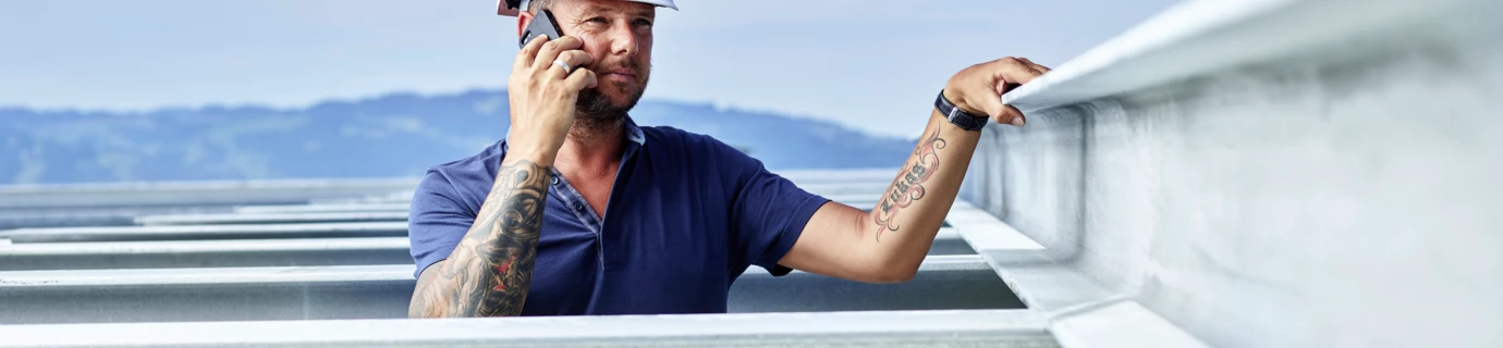 Ein Mann mit Helm und Rhomberg-Logo telefoniert und lehnt an einem Träger unter blauem Himmel (Gleisen).