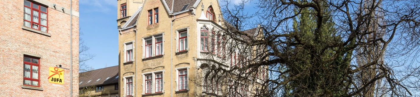 Blick auf das Benger Areal mit einem gelben Gebäude, grünen Wiesen und einem großen kahlen Baum im Vordergrund.
