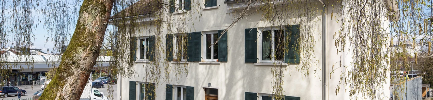 Aussenansicht des Mehrfamilienhauses Klostergasse 30 in Dornbirn mit einem blühenden Baum im Vordergrund.