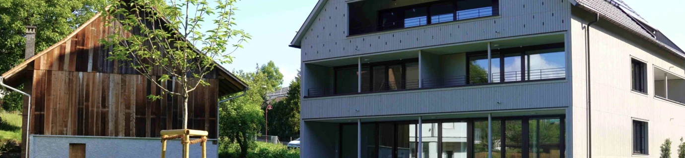 Modernes Mehrfamilienhaus mit Balkonen und Solarpanelen auf dem Dach neben einem traditionellen Holzhaus mit Gründach und Wiese (Götzis, Außenanlage)