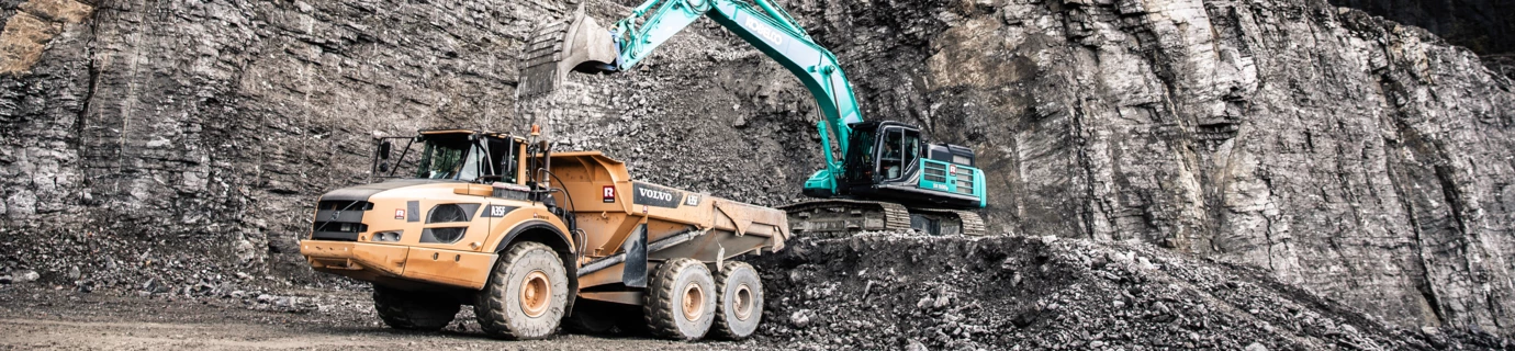 Ein türkiser Bagger mit Greifarm kippt Schotter in einen gelben Volvo Lkw in einem Steinbruch, im Hintergrund ist eine steile Felswand zu sehen (Ressourcen Center Rheintal).