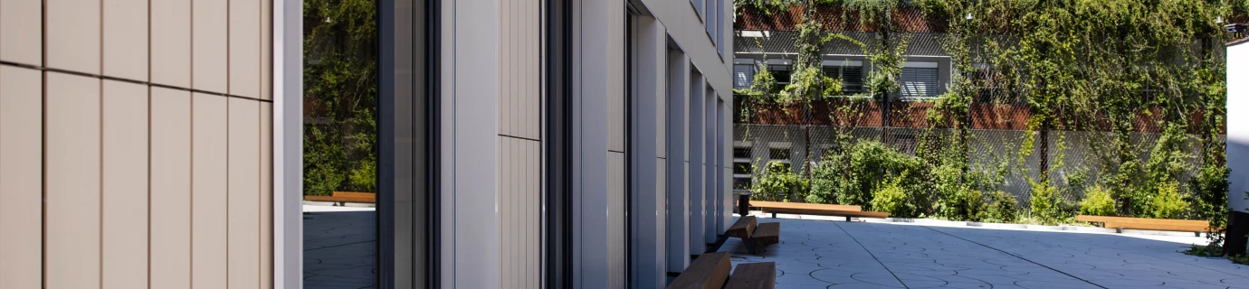 Seitliche Ansicht eines modernen Gebäudes mit Glasfenstern und beigefarbenen Paneelen, daneben eine Terrasse mit Holzbänken und einem begrünten Haus im Hintergrund (Baerahus, Feldkirch).