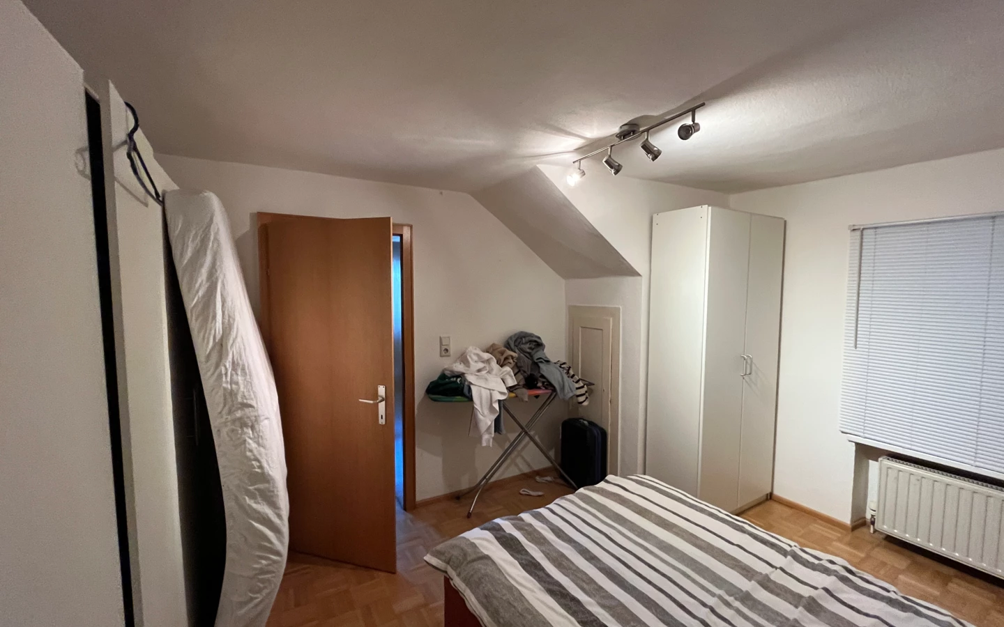 Unordentliches Schlafzimmer mit gestreiftem Bett, Bügelbrett voller Kleidung, Matratze an der Wand und Kleiderschrank.