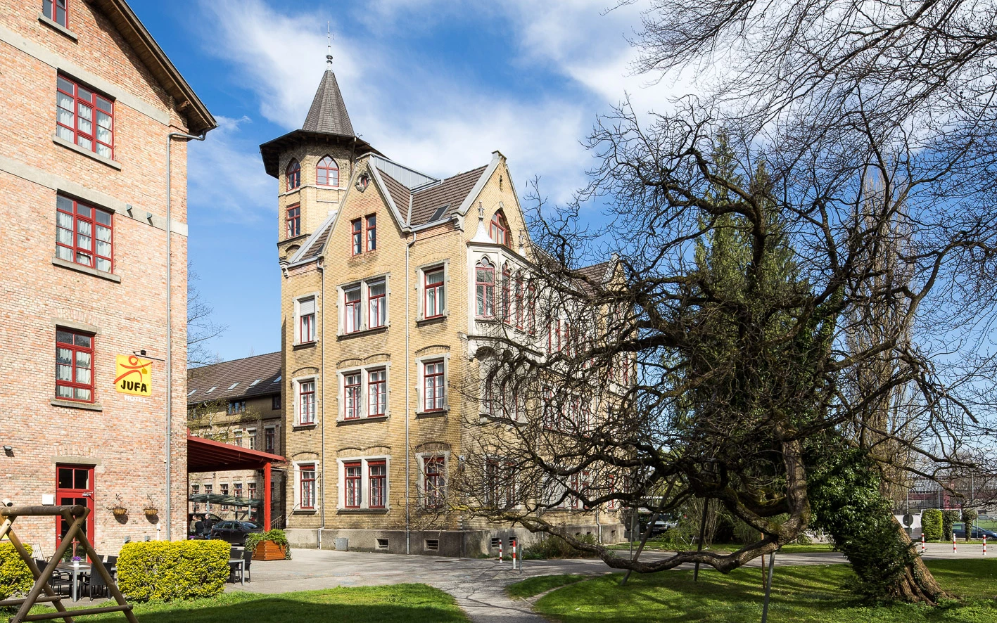 Blick auf das Benger Areal mit einem gelben Gebäude, grünen Wiesen und einem großen kahlen Baum im Vordergrund.