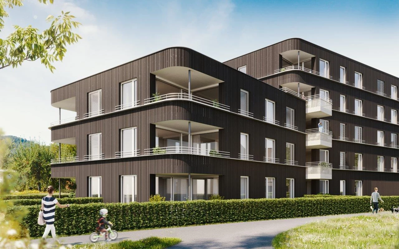 Modernes, mehrstöckiges Wohngebäude mit dunkler Fassade und Balkonen. Eine Frau geht mit einem Kind auf dem Laufrad vorbei, ein Mann spaziert mit Hunden (Rheindorf).