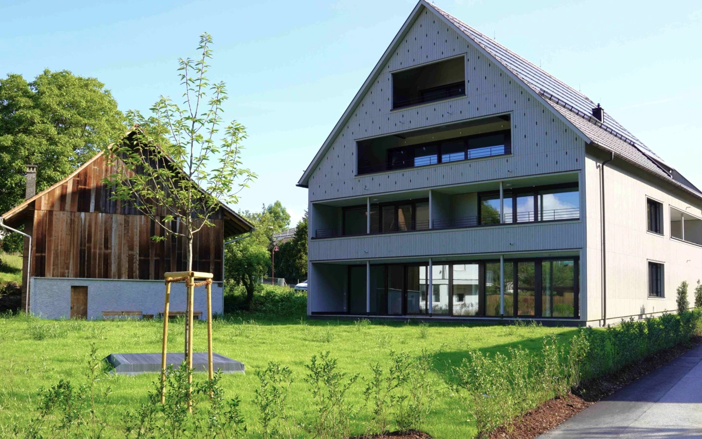 Modernes Mehrfamilienhaus mit Balkonen und Solarpanelen auf dem Dach neben einem traditionellen Holzhaus mit Gründach und Wiese (Götzis, Außenanlage)