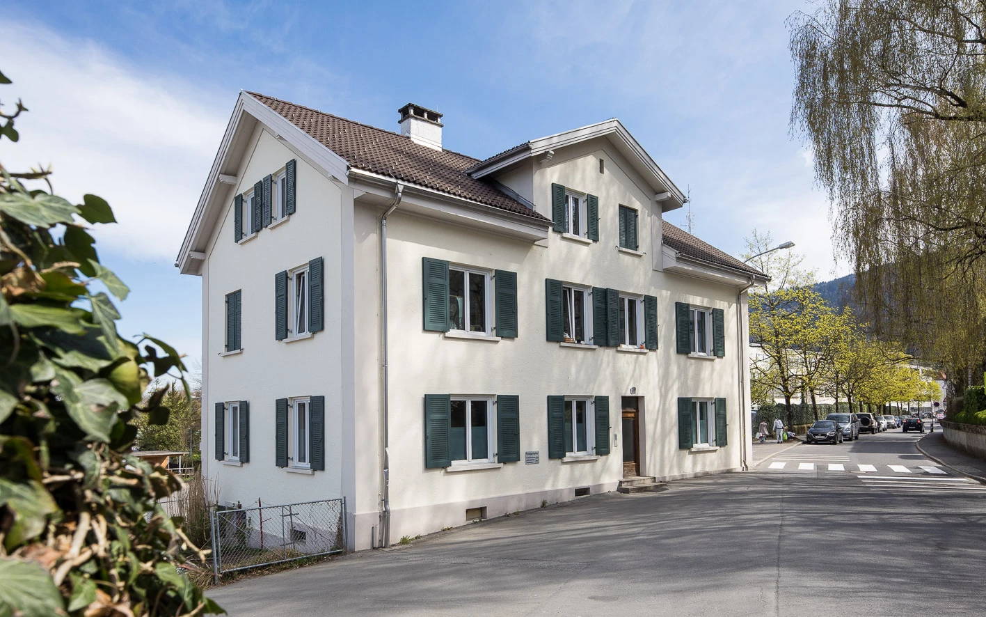 Weißes dreistöckiges Gebäude an der Klostergasse mit grünen Fensterläden und einem hellbraunen Ziegeldach. Eine Straße und parkende Autos sind im Vordergrund zu sehen.