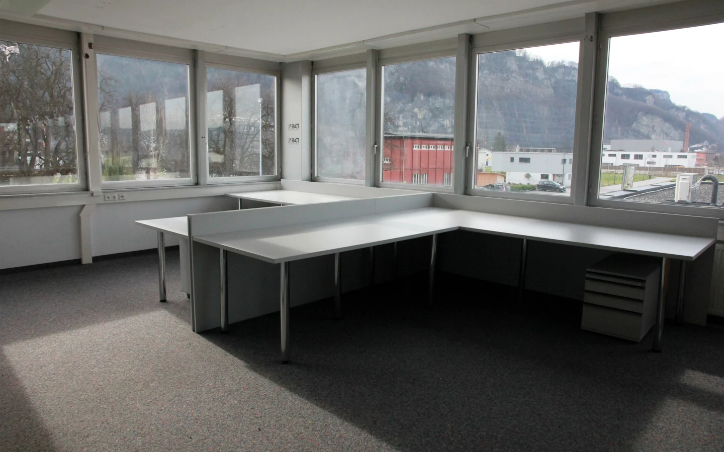 Leeres Eckbüro im Froschweg mit langen Schreibtischen, Fenstern mit Bergblick und Spannteppichboden (Büro).