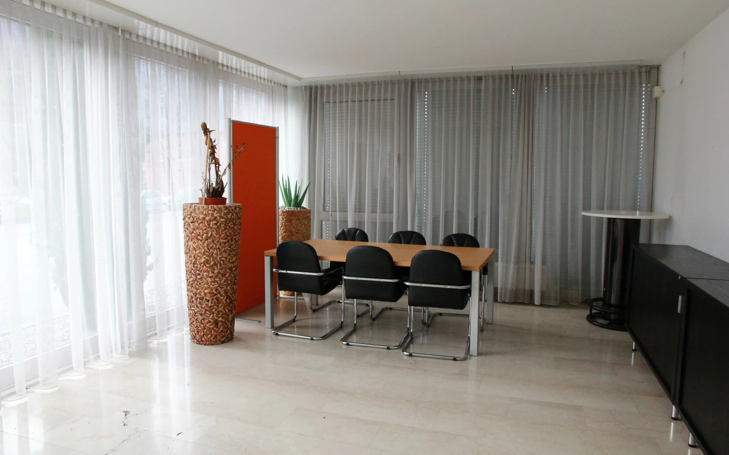 Ein modernes Büro oder Besprechungsraum mit einem Holztisch, sechs schwarzen Stühlen und zwei beigen Pflanzgefäßen mit Pflanzen vor Fenstern mit weißen Vorhängen (Froschweg).