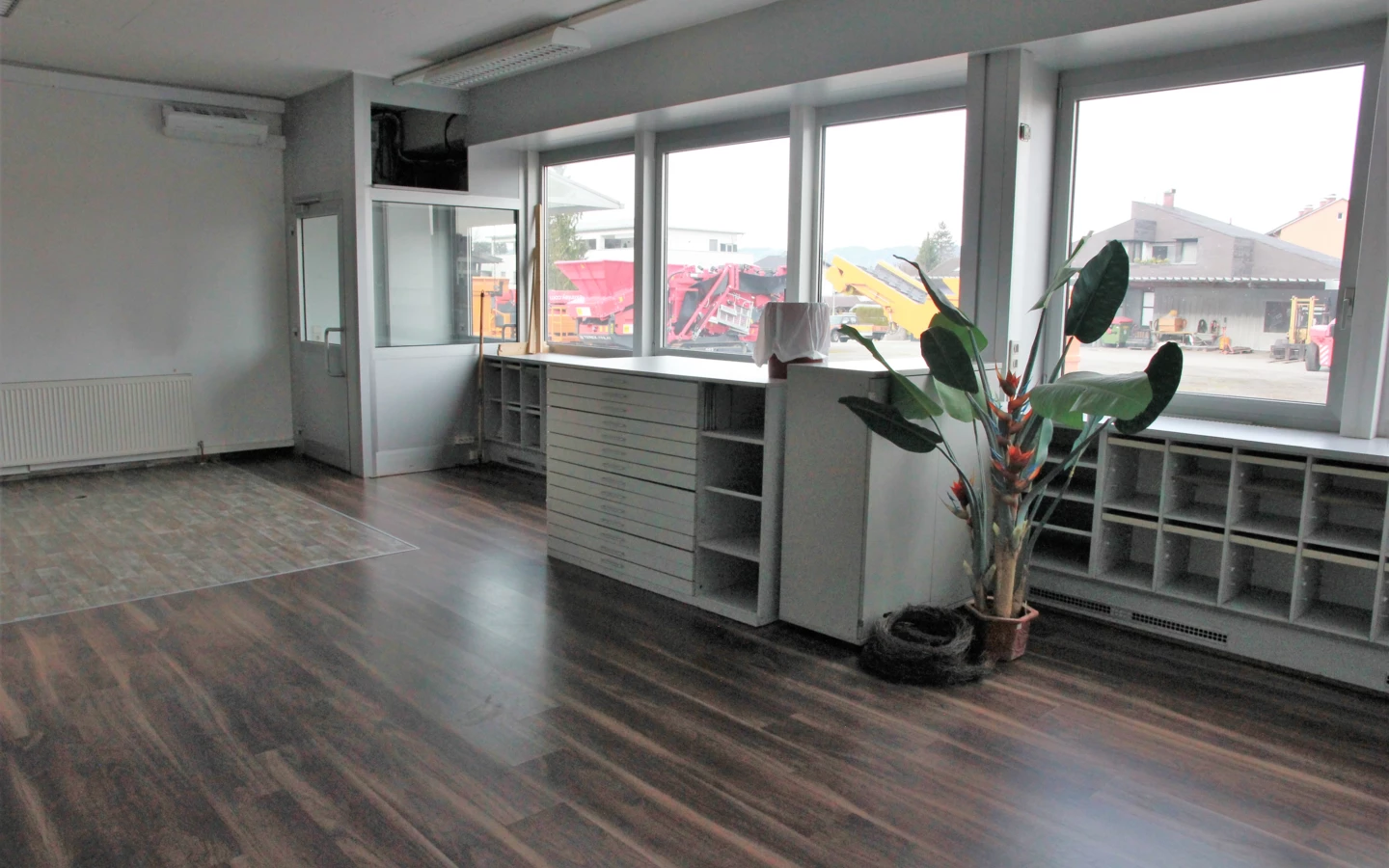 Leeres Büro im Froschweg mit Holzboden, großen Fenstern und Blick auf Baufahrzeuge, mit einer Pflanze und einem Schrank.