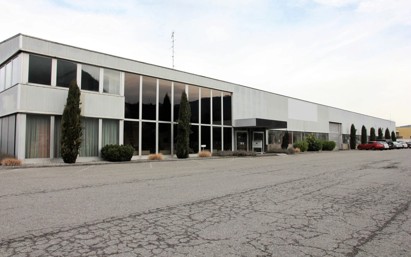 Langes, modernes Bürogebäude und Lagerhalle mit großen Fenstern an einem bewölkten Tag, davor ein Parkplatz mit Autos (Froschweg).
