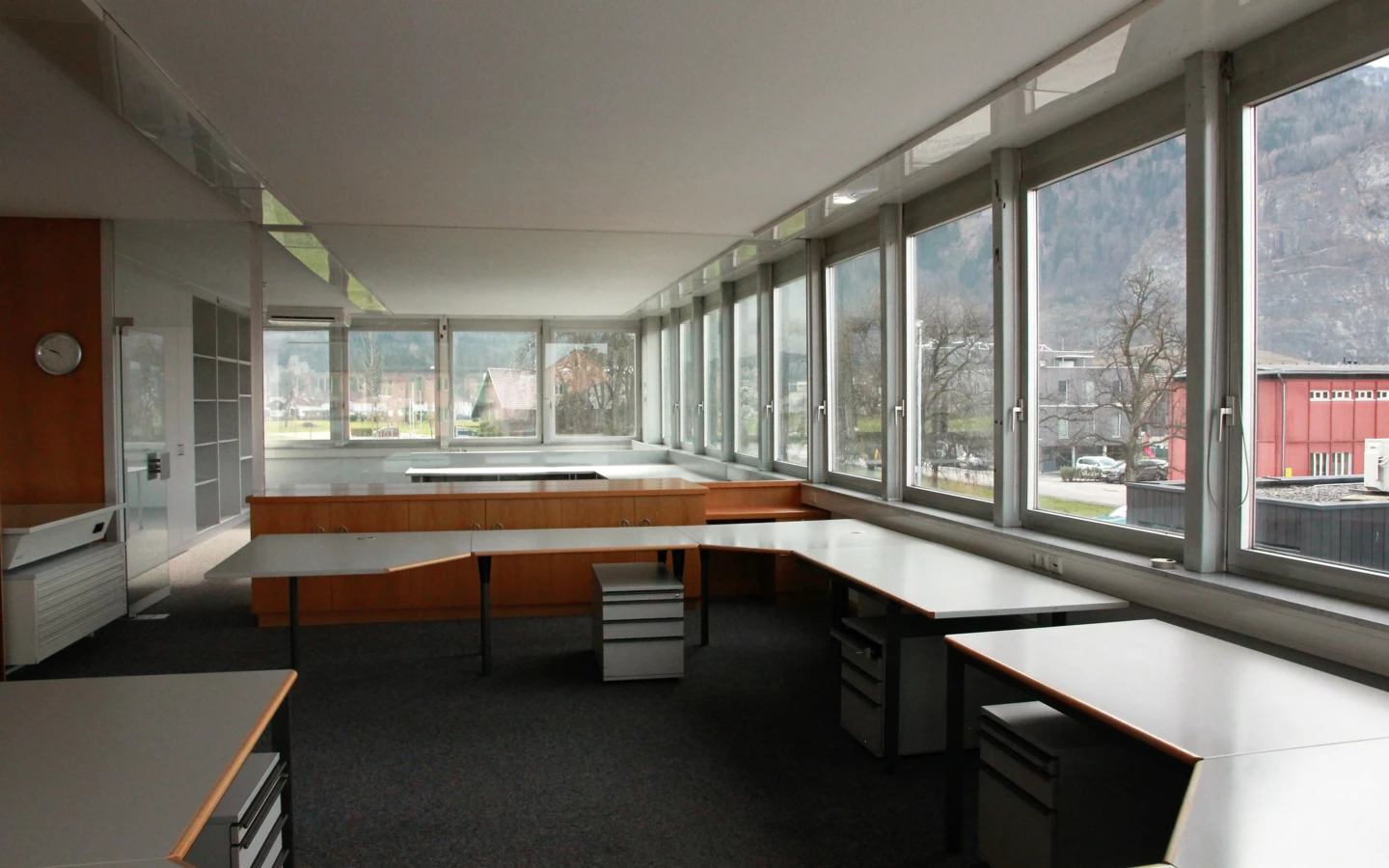 Modernes Büro mit großen Fenstern mit Blick auf Berge, leeren Schreibtischen und Rollcontainern in einer offenen Bürolandschaft (Froschweg).