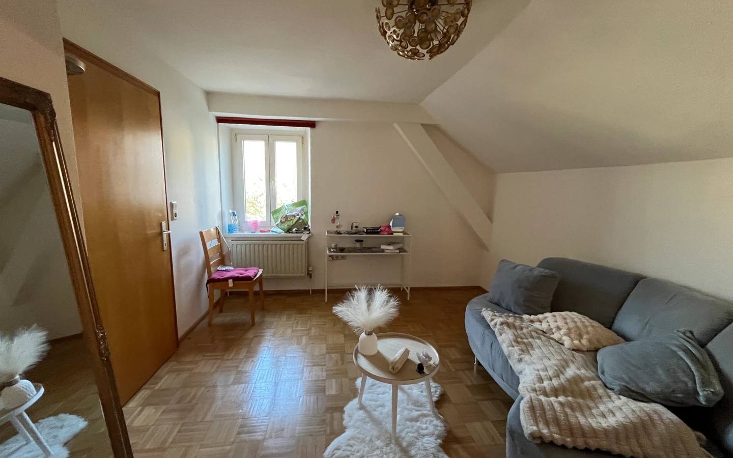 Wohnzimmer mit grauem Sofa und Decke, Beistelltisch auf Schaffell-Teppich, Holzboden, Fenster und Holztür.