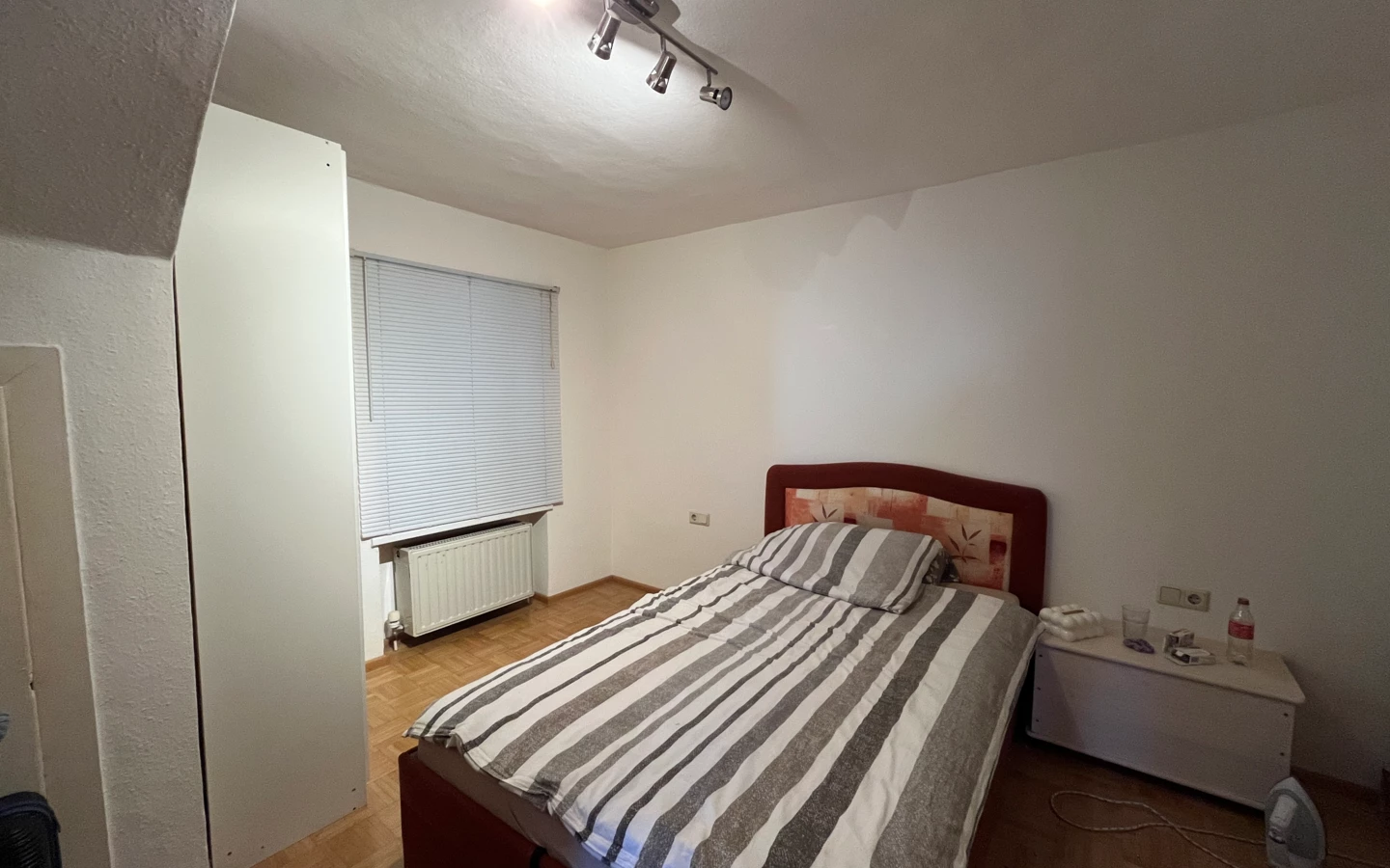 Ein bescheidenes Schlafzimmer mit Ehebett, brauner Bettwäsche und gestreifter Decke. Ein weißer Nachttisch steht daneben.
