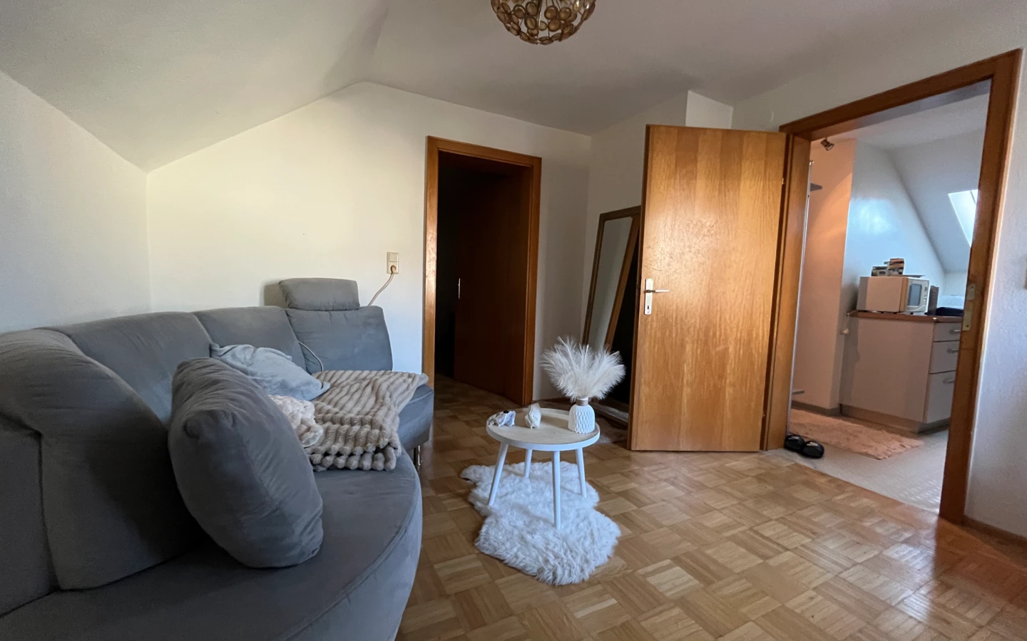 Ein Zimmer mit einer grauen Couch, einem runden Beistelltisch auf einem Kunstfellteppich und zwei offenstehenden Holztüren.