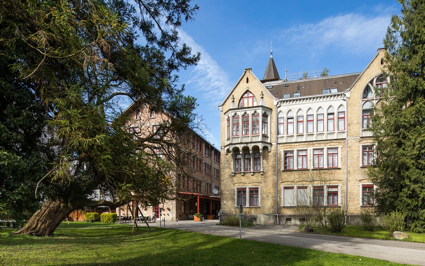 Zwei historische Gebäude am Benger Areal mit einem alten Baum im Vordergrund und blauem Himmel