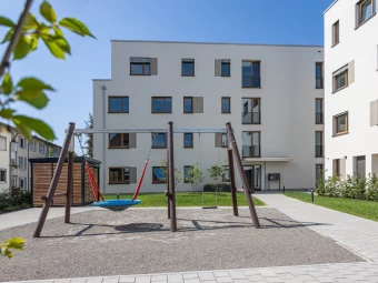 Spielplatz mit Nestschaukel und traditioneller Schaukel vor einer modernen Wohnanlage mit weißen Fassaden und großen Fenstern (Innenbereich)
