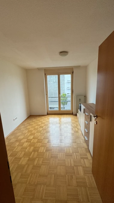 Leeres, helles Zimmer mit Parkettboden, einem Fenster mit Jalousien und einem schmalen Schrank an der Seite.