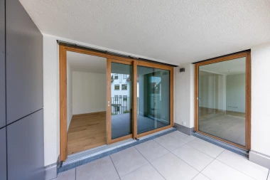 Ein moderner Balkon mit hellgrauen Fliesen und großen Holz-Glasschiebetüren, die in eine leere Wohnung mit Holzboden führen (Hüni, Balkonansicht).