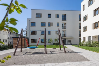 Spielplatz mit Nestschaukel und traditioneller Schaukel vor einer modernen Wohnanlage mit weißen Fassaden und großen Fenstern (Innenbereich)