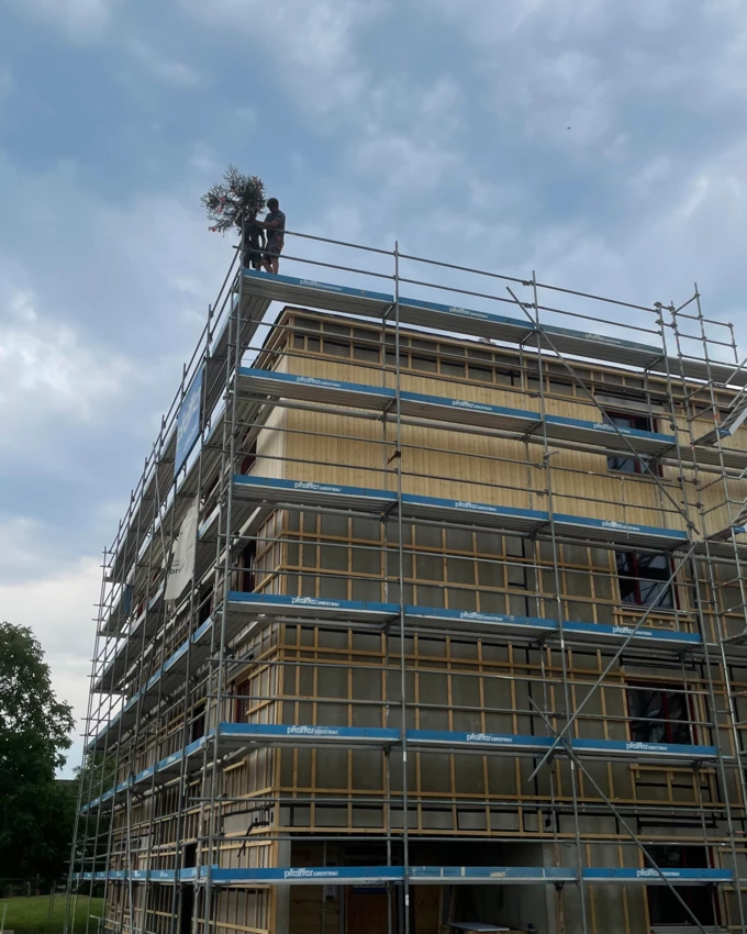 Holz auf Holz: Zwei Zimmerer von Sutter Holzbau beim Stellen des Firstbaums für den Neubau in der Neubaugasse, Lauterach. Darunter ist die Holzfassade sehr gut zu erkennen, die aktuell montiert wird.

