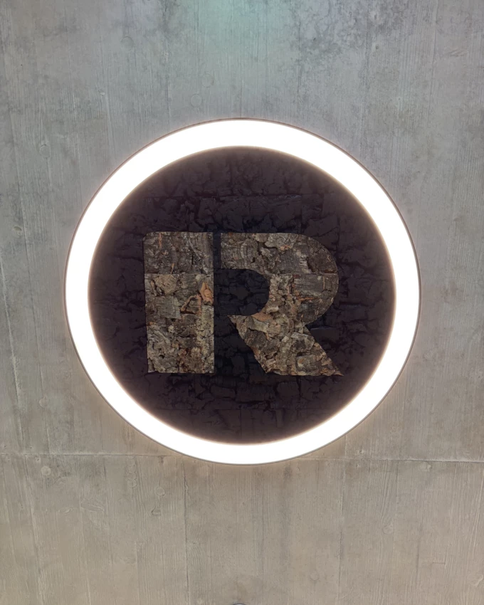 Rhomberg R-Logo aus Kork, in einer kreisförmigen Leuchte mit Umrandung aus LED-Licht an einer grauen Betonwand (Nachhaltigkeit).
