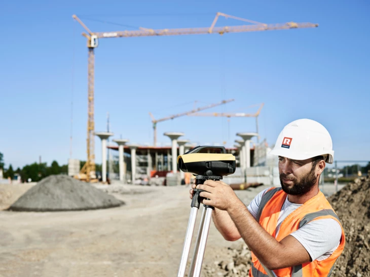 Tiefbau Mann vermisst Baustelle mit speziellem Werkzeug und Kran im Hintergrund Tiefbau Mann vermisst Baustelle mit speziellem Werkzeug und Kran im Hintergrund
