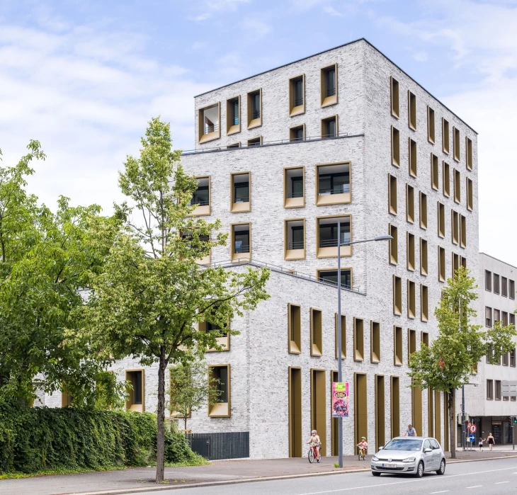 Modernes Mehrfamilienhaus mit heller Backsteinfassade und goldfarbenen Fensterrahmen unter blauem Himmel mit Bäumen (Kreuzgasse).