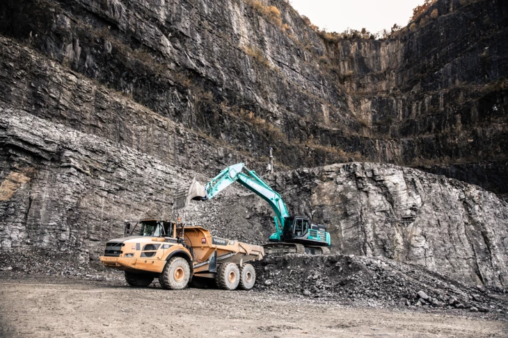 Ein türkiser Bagger mit Greifarm kippt Schotter in einen gelben Volvo Lkw in einem Steinbruch, im Hintergrund ist eine steile Felswand zu sehen (Ressourcen Center Rheintal).