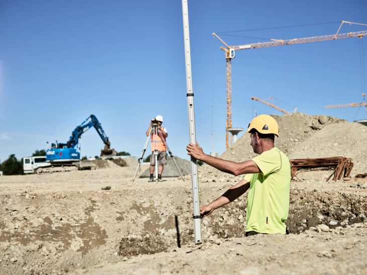 Tiefbau Mann hält Metallstange in eine Grube, anderer Mann vermiss im Hintergrund