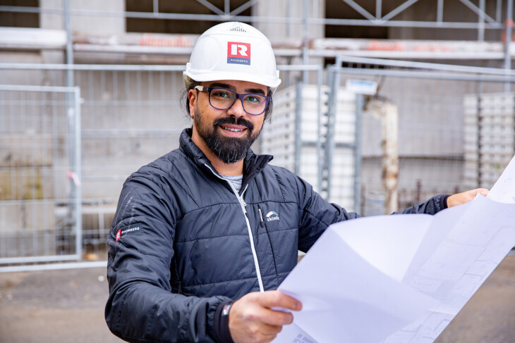Goekhan Uelker hält Plan vor sich auf einer Baustelle und lächelt in die Kamera Goekhan Uelker hält Plan vor sich auf einer Baustelle und lächelt in die Kamera