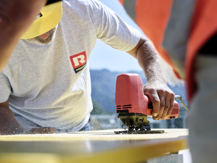 Hochbau Rhomberg Bau Mann schneidet Brett mit Stichsäge zu