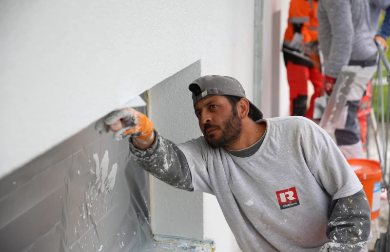 Arbeiter in Schutzkleidung verputzt eine Fensterlaibung an einem Gebäude in Meiningen, verschmutzte Bauwand (Verputzarbeiten, Hochbau)