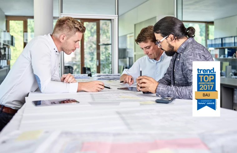 Drei Bauingenieure besprechen Baupläne und arbeiten mit einem Tablet. In der oberen rechten Ecke ist das Logo „trend. TOP ARBEITGEBER 2017 BAU“ zu sehen (Bester).