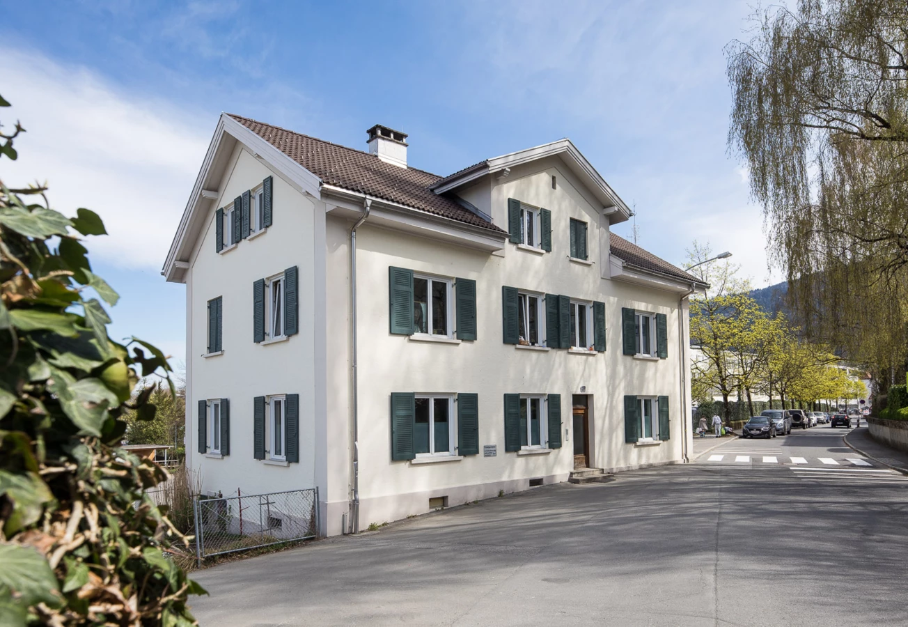 Weißes dreistöckiges Gebäude an der Klostergasse mit grünen Fensterläden und einem hellbraunen Ziegeldach. Eine Straße und parkende Autos sind im Vordergrund zu sehen.
