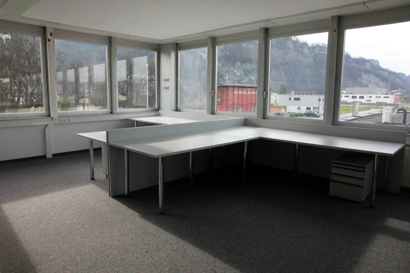 Leeres Eckbüro im Froschweg mit langen Schreibtischen, Fenstern mit Bergblick und Spannteppichboden (Büro).