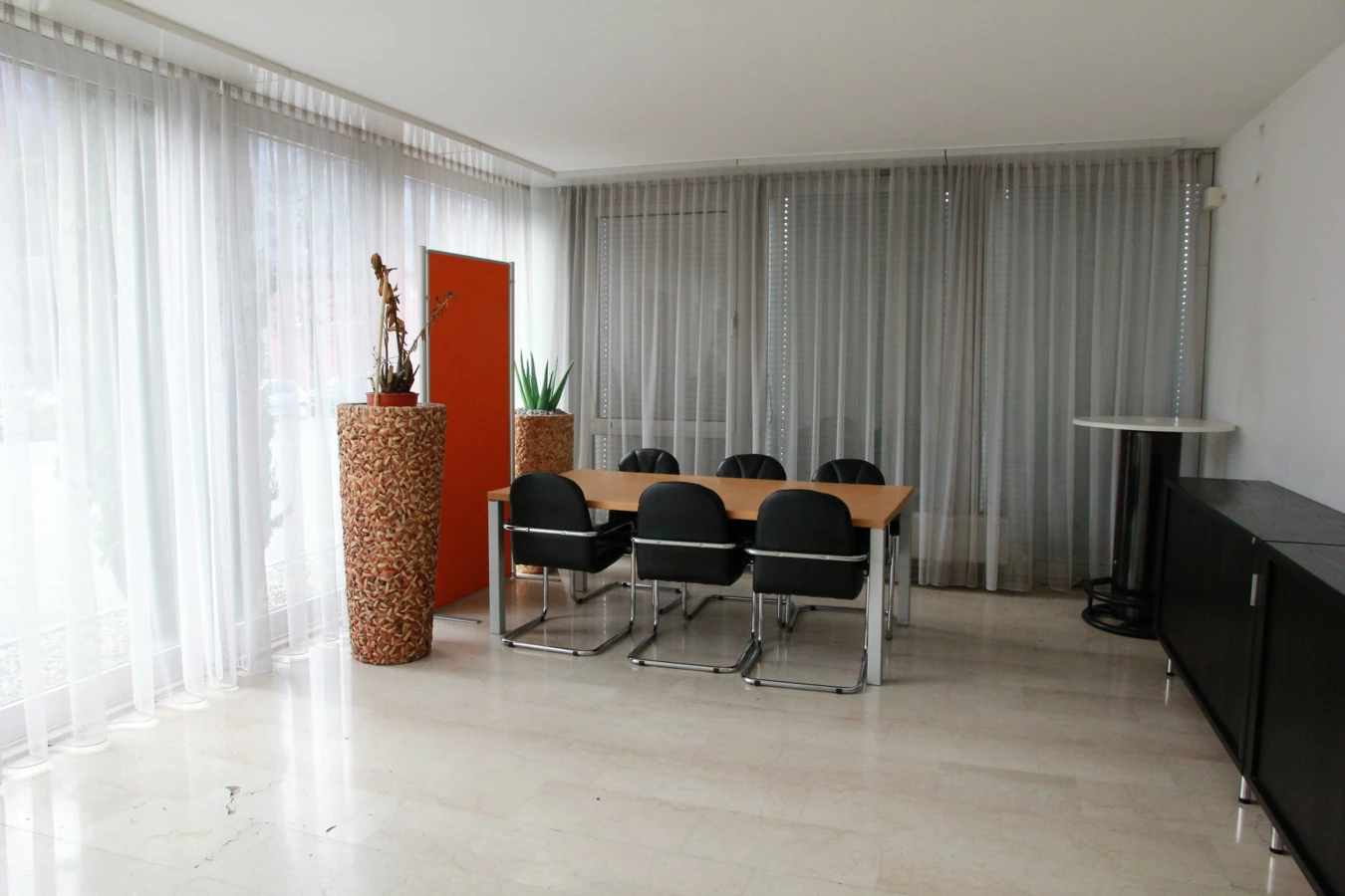 Ein modernes Büro oder Besprechungsraum mit einem Holztisch, sechs schwarzen Stühlen und zwei beigen Pflanzgefäßen mit Pflanzen vor Fenstern mit weißen Vorhängen (Froschweg).