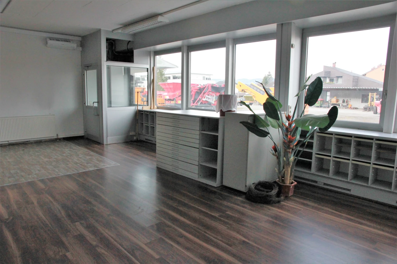 Leeres Büro im Froschweg mit Holzboden, großen Fenstern und Blick auf Baufahrzeuge, mit einer Pflanze und einem Schrank.