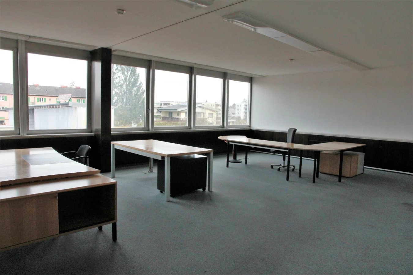 Leeres Büro mit Schreibtischen, Stühlen und einer Fensterfront, die eine Ansicht auf Gebäude und Bäume bietet (Froschweg).