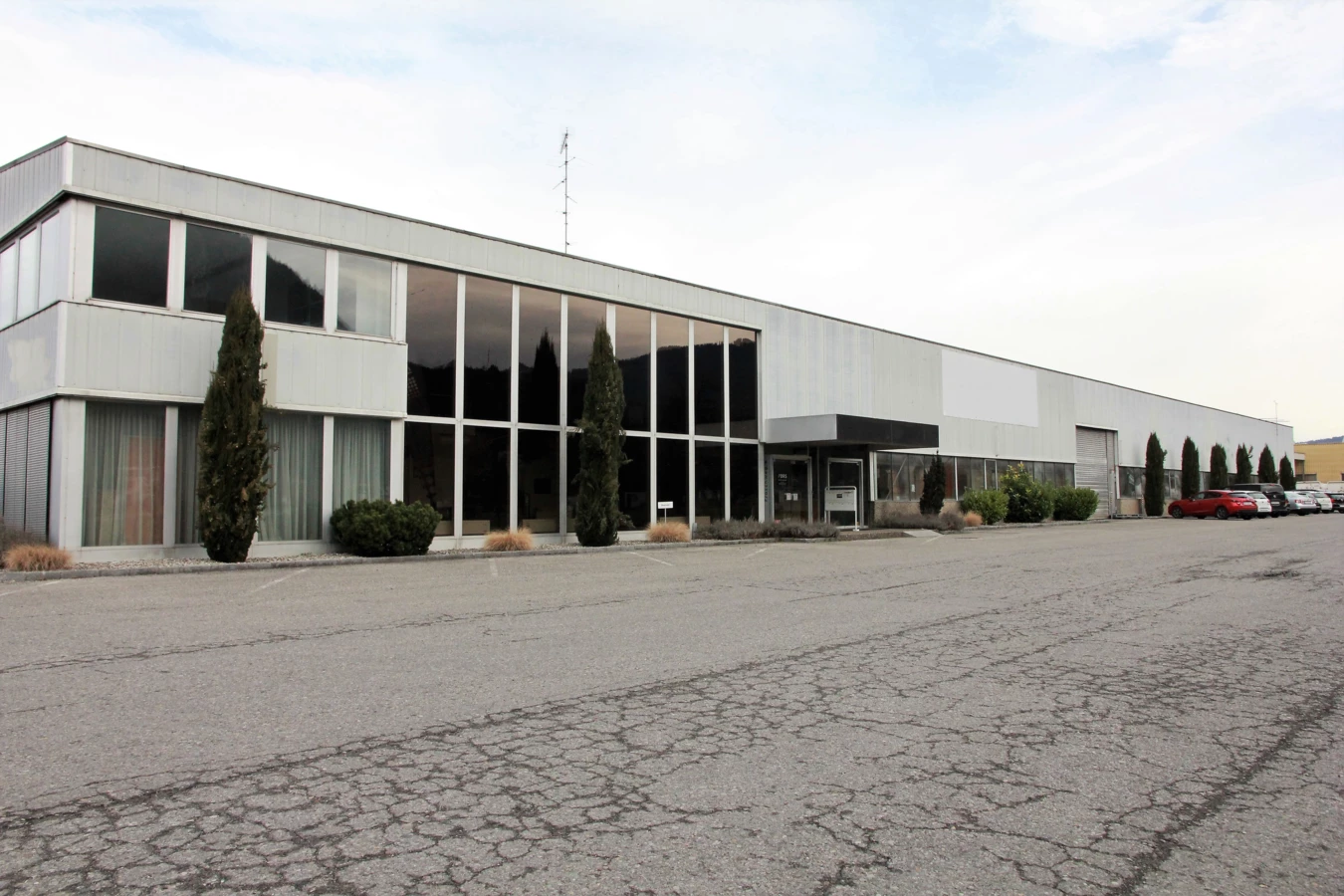 Langes, modernes Bürogebäude und Lagerhalle mit großen Fenstern an einem bewölkten Tag, davor ein Parkplatz mit Autos (Froschweg).