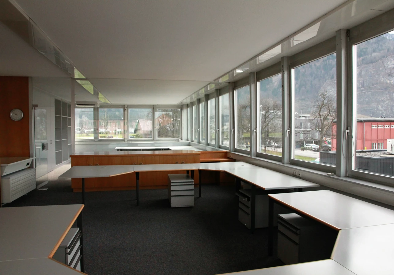 Modernes Büro mit großen Fenstern mit Blick auf Berge, leeren Schreibtischen und Rollcontainern in einer offenen Bürolandschaft (Froschweg).